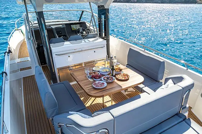 Luxury boat interior featuring a dining table with food, drinks, and grey seating, overlooking sparkling blue water.